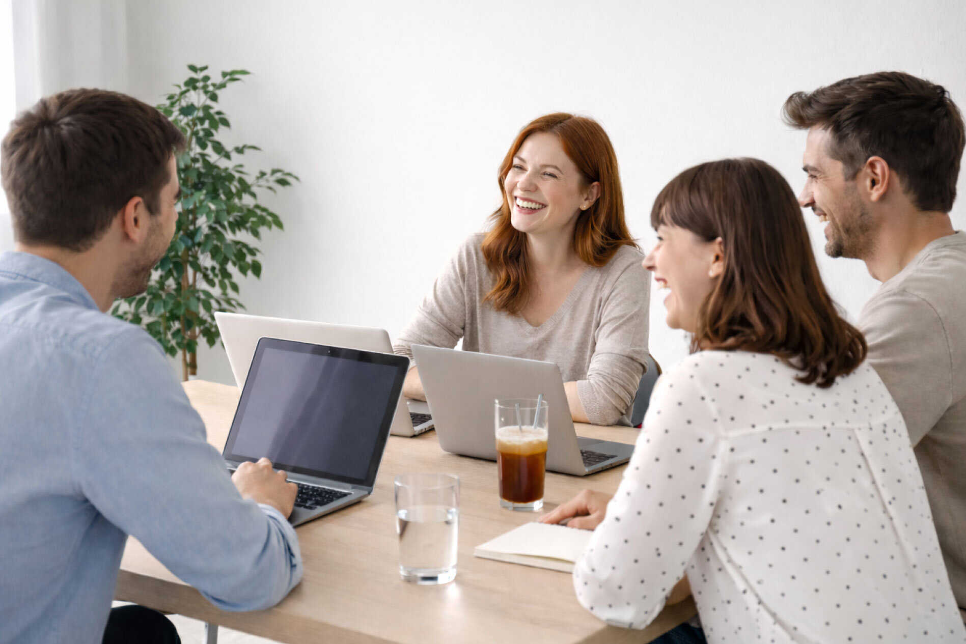 KI-generiertes Bild: Vier Personen sitzen an einem Tisch in einem modernen Büro und führen ein Gespräch. Auf dem Tisch stehen Laptops, Notizblöcke und ein Glas mit Getränk.