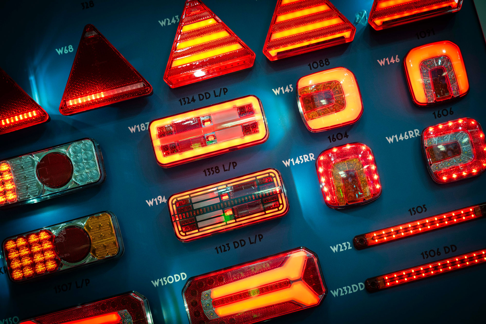 Verschiedene leuchtende Fahrzeug- und Anhänger­rückleuchten in unterschiedlichen Formen und Größen, angeordnet auf einer blauen Präsentationsfläche.
